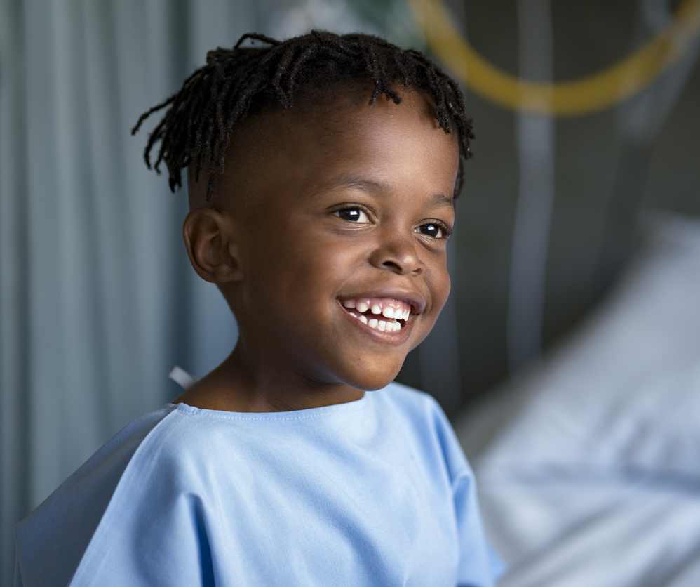 Smiling boy looking away sitting on bed at ward. Cute male patient is in blue gown. He is at hospital.